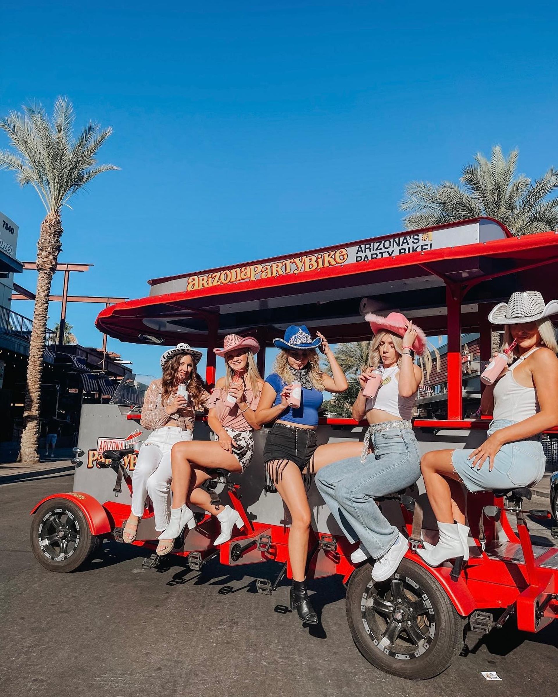 Five women in cowboy hats and boots laughing and sipping drinks on a red open-air party bike under palm trees on a sunny Arizona street.