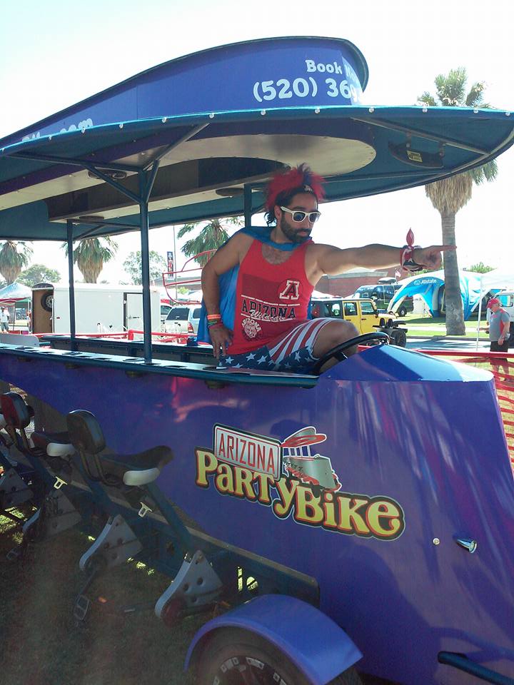 Bearded man in a red tank top and American-flag shorts striking a pose and pointing from a purple party-bike pedal bar under a blue canopy at an outdoor Arizona festival.