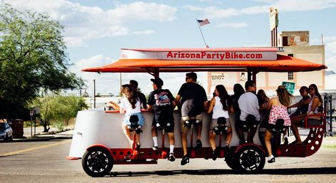 Group of young adults pedaling a red open-air party bike down a sunny Arizona street, laughing under a blue sky with an American flag on top.