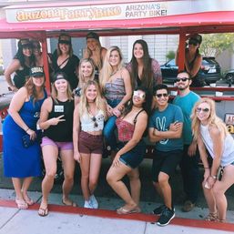 Large group of smiling young adults posing on and beside a red open-air party bike on a sunny Arizona street, wearing casual summer clothes and sunglasses.