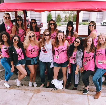 Cheerful bachelorette crew of women in matching pink tank tops and sunglasses, one wearing a white sash, posing under a red party cart on a downtown street.