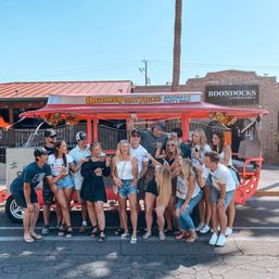 Cheerful group of friends posing beside a bright red open-air party bike on a sunny Arizona street by a brick patio restaurant