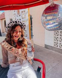 Smiling bride-to-be wearing a leopard cowgirl hat and glitter sash, holding a 'Bride' can cooler next to an iridescent balloon on a sunny sidewalk ride