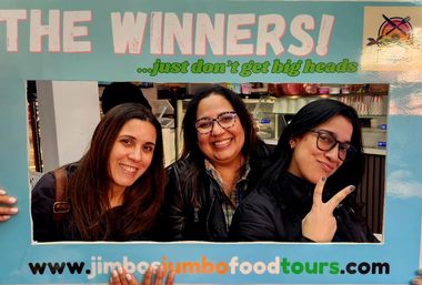 Three smiling women pose behind a large blue photo frame that reads THE WINNERS with a playful green tagline, inside an indoor food market — one woman flashes a peace sign.
