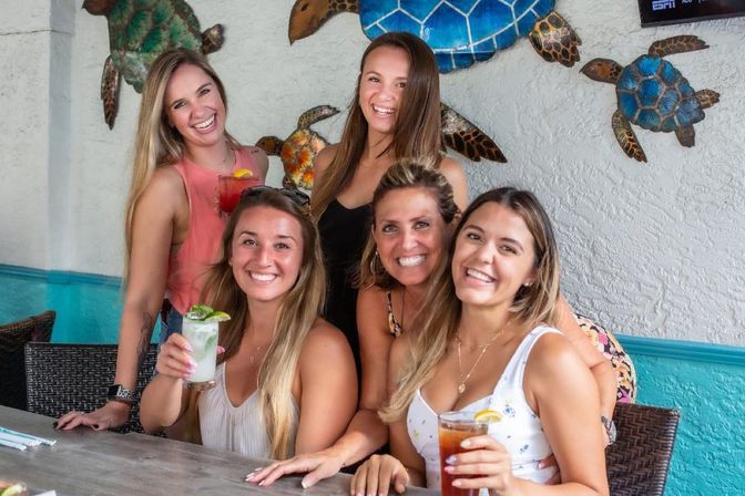 Five smiling women enjoying cocktails at a colorful tropical beach bar with decorative sea turtle wall art