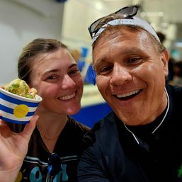 Bright selfie of two smiling people holding a striped cup of colorful ice cream inside a lively ice cream shop