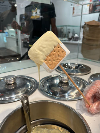 White chocolate–dipped s'mores pop (graham cracker and marshmallow) on a skewer dripping over a dessert shop dipping station