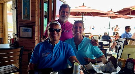 Three smiling people in colorful polo shirts seated at a beachside restaurant patio with umbrellas and sunlit ocean in the background, table with drinks and snacks.