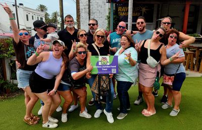 Playful group of adults posing on an outdoor patio for a Tampa Bay food tour, many making peace signs and holding a colorful tour sign