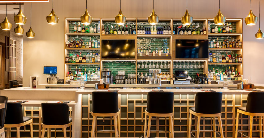Sleek upscale cocktail bar interior with glowing backlit liquor shelves, green subway-tile backsplash, gold pendant lights, a long marble counter and black leather-topped wooden bar stools.