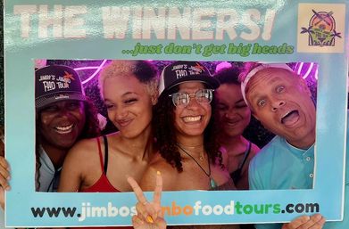 Five people smiling and laughing inside a turquoise “THE WINNERS” photo-frame at a neon-lit food tour photo booth, two wearing caps and one flashing a peace sign