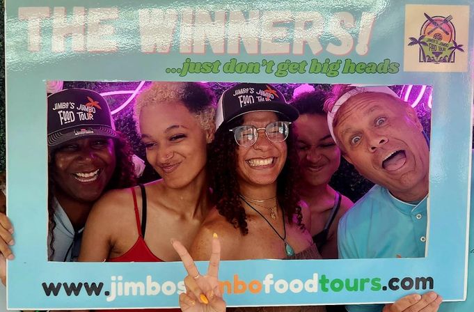 Five people smiling and laughing inside a turquoise “THE WINNERS” photo-frame at a neon-lit food tour photo booth, two wearing caps and one flashing a peace sign