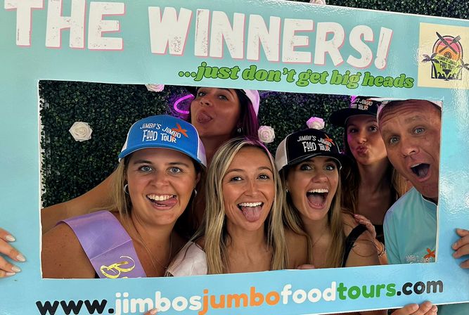 Six adults making playful faces and sticking out tongues inside a pastel "The Winners" photo-frame at an outdoor food‑tour event, wearing caps and a sash against a green foliage backdrop with white roses.
