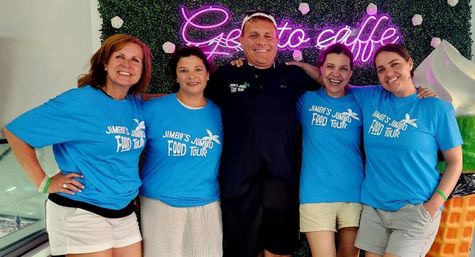 Smiling group of five adults — four women in matching bright-blue food-tour shirts and a man in a black polo — posing inside a gelato café in front of a green floral wall and pink neon sign.