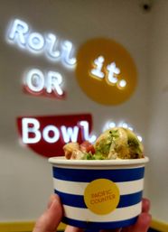 Hand holding a blue-and-white striped cup with a poke-style bowl topped with avocado, diced fish and creamy sauce, blurred neon wall sign and colorful fast-casual interior in the background
