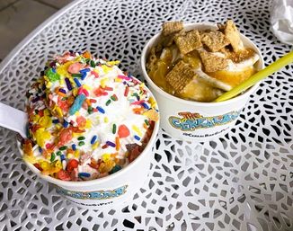 Two cereal-topped soft-serve dessert cups — one with whipped topping, rainbow sprinkles and fruity cereal, the other with caramel drizzle and square cinnamon cereal — on a white decorative metal patio table.