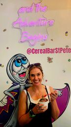 Smiling woman holding a cup of cereal topped with sprinkles and chocolate, posing in front of a colorful cereal-shop mural and pink neon sign reading 'and the Adventure Begins'.