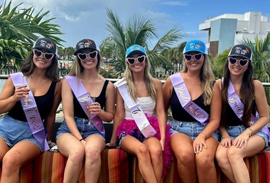 Five smiling friends on a sunny seaside rooftop celebrating a bachelorette — center bride in a white sash and pink feather skirt, all wearing heart-shaped sunglasses, matching caps and purple sashes, denim shorts and drinks, with palm trees and coastal buildings in the background.
