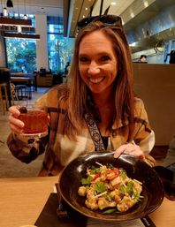 Smiling woman in a plaid jacket raises a cherry‑garnished cocktail while presenting a black bowl of roasted cauliflower topped with sliced red chili, cilantro and scallions in a modern urban restaurant with large windows and warm lighting.