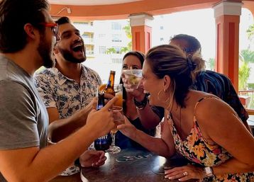 Group of friends laughing and toasting beers and cocktails at a sunny open-air bar patio with palm trees and resort-style buildings in the background.