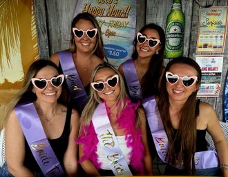 Smiling group of five friends at a tropical beach-bar bachelorette party, wearing purple sashes and pink heart-shaped sunglasses; bride in center with white sash and pink feather boa posing in front of rustic wooden signage