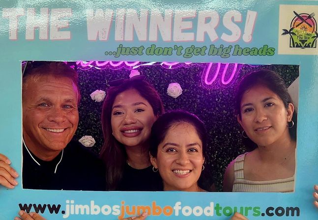 Four smiling adults posing behind a pastel-blue "The Winners!" photo frame at a food-tour event, with a green floral wall, white roses and a neon sign backdrop.