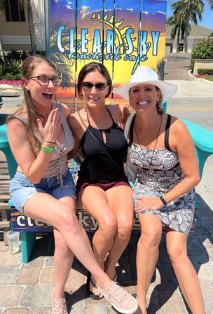 Three smiling women seated on a turquoise oversized chair in a sunny beach town, posed in front of a colorful beachside-cafe sunset sign, wearing summer outfits, sunglasses and a white sun hat