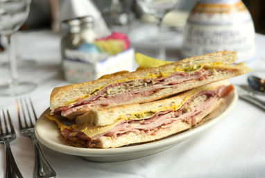 Close-up of a pressed Cuban-style sandwich with layers of ham, roast pork, melted cheese, pickles and mustard on toasted crusty bread, served on a white plate with a pickle spear and blurred restaurant table setting