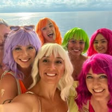Smiling group selfie of friends wearing neon wigs (purple, blonde, orange, green, pink) on a boat with calm ocean and horizon in the background