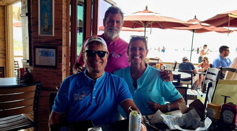 Three smiling people in colorful polo shirts seated at a beachside restaurant patio with umbrellas and sunlit ocean in the background, table with drinks and snacks.