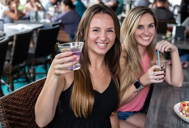 Two friends smiling and toasting cocktails at a lively restaurant patio bar with a casual brunch crowd in the background.