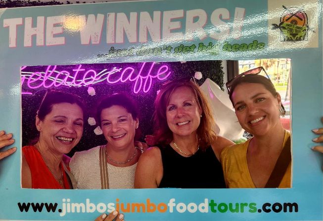 Four smiling women posing behind a light-blue “The Winners” photo-frame prop in front of a neon “gelato caffe” sign at a colorful food-tour event