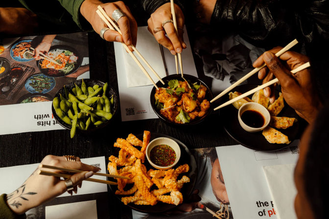 Overhead shot of hands with chopsticks sharing Asian small plates at a restaurant — bowl of edamame, gyoza with soy dip, crispy battered seafood sticks with dipping sauce, and seasoned chicken garnished with cilantro on a dark table.