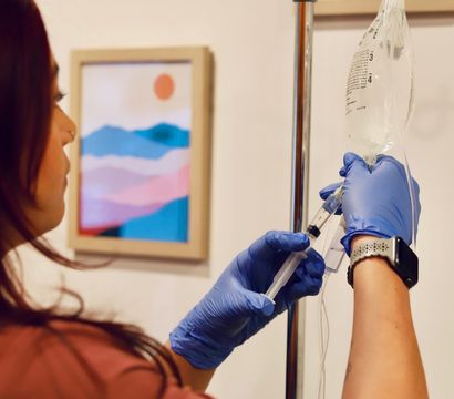 Healthcare worker in blue gloves prepping an IV drip bag and syringe on a chrome IV pole in a bright treatment room with colorful abstract wall art