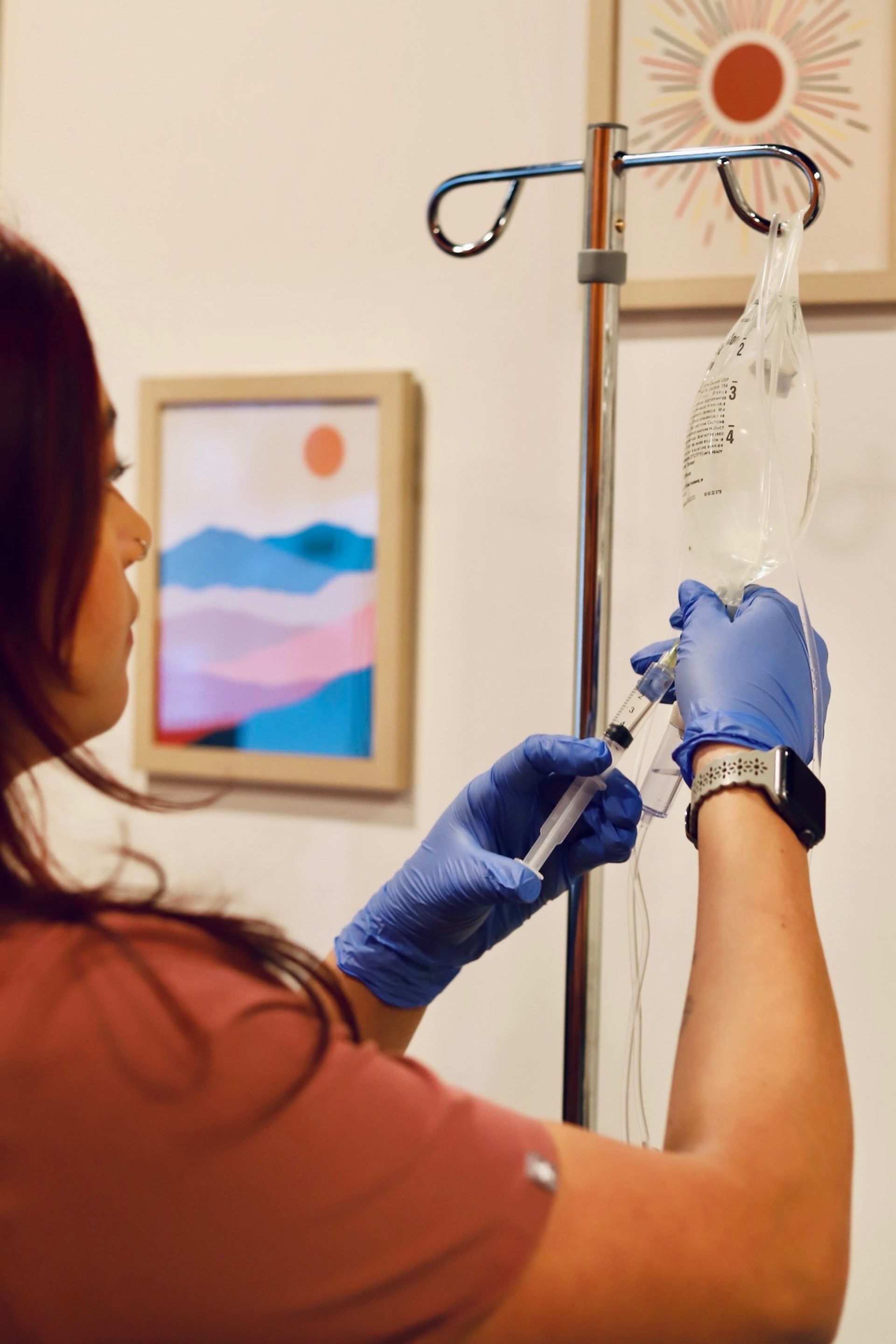 Healthcare worker in blue gloves prepping an IV drip bag and syringe on a chrome IV pole in a bright treatment room with colorful abstract wall art