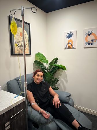 Smiling woman reclining in a clinic treatment chair receiving a yellow IV infusion, cozy wellness room with potted plant and modern framed wall art