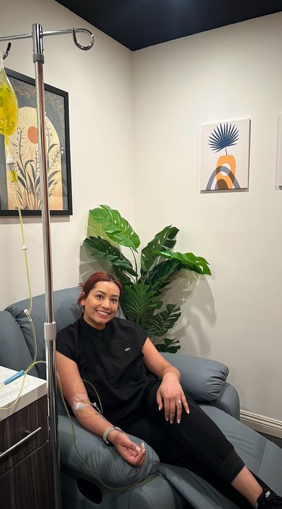Smiling woman reclining in a clinic treatment chair receiving a yellow IV infusion, cozy wellness room with potted plant and modern framed wall art