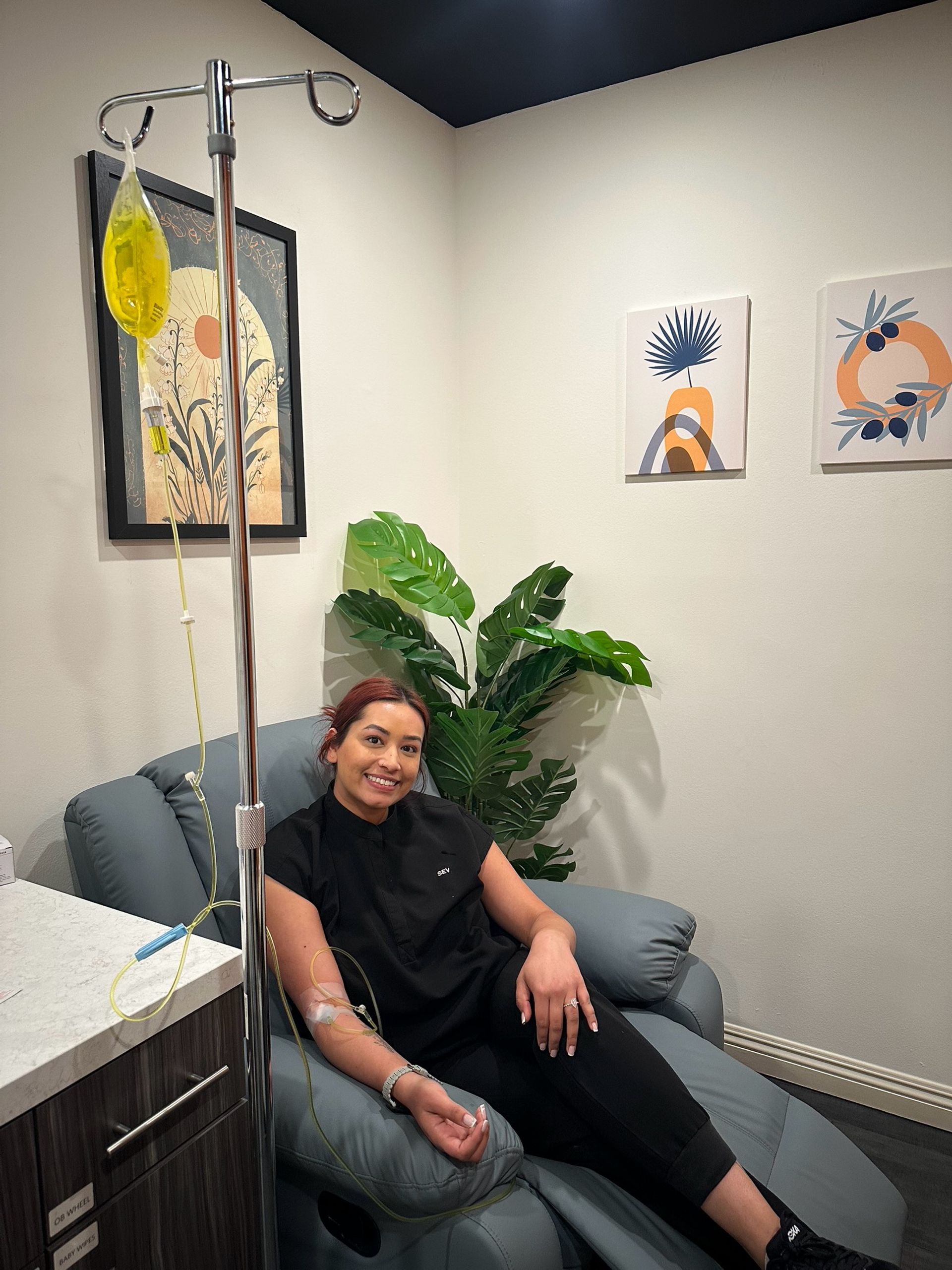Smiling woman reclining in a clinic treatment chair receiving a yellow IV infusion, cozy wellness room with potted plant and modern framed wall art