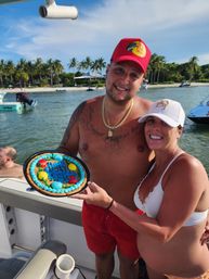 Smiling couple celebrating on a boat holding a colorful cookie cake that says 'Happy Birthday', with a tropical palm-lined beach and anchored boats in the background.