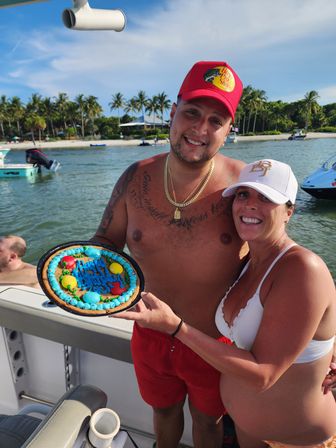 Smiling couple celebrating on a boat holding a colorful cookie cake that says 'Happy Birthday', with a tropical palm-lined beach and anchored boats in the background.