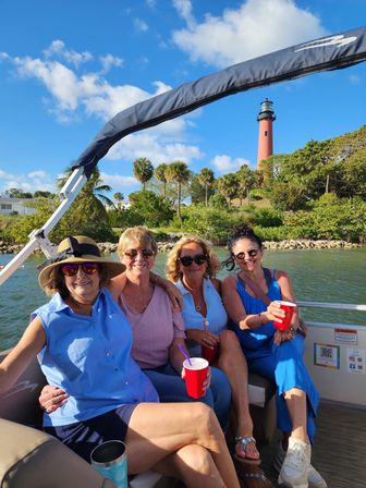 Four women smiling and relaxing on a boat holding red cups, sunny coastal scene with palm trees and a tall red lighthouse rising above the shore under a bright blue sky.