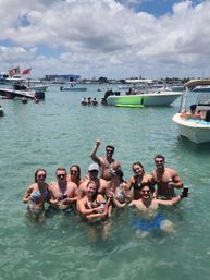 Smiling group of friends waist‑deep in clear turquoise bay holding drinks, surrounded by anchored boats and a sunny, partly cloudy sky