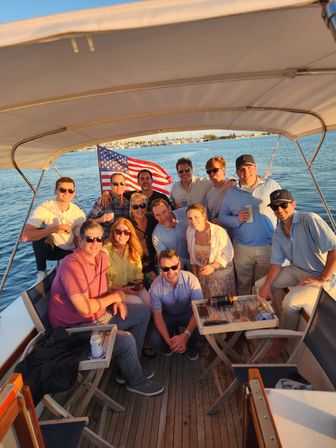 Sunlit group of friends on a leisure boat cruise at sunset, gathered on the teak deck with drinks and an American flag waving, marina and calm harbor water in the background.