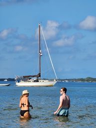 Two people wading in shallow turquoise water near an anchored sailboat under a bright blue sky with puffy clouds and a palm-lined shoreline