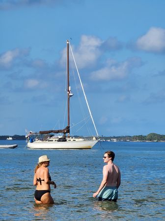 Two people wading in shallow turquoise water near an anchored sailboat under a bright blue sky with puffy clouds and a palm-lined shoreline