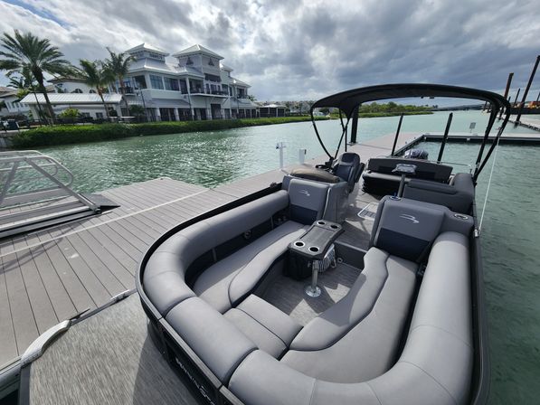 Spacious gray pontoon boat with wraparound cushioned seating and Bimini top docked at a palm-lined waterfront marina by a white coastal building and turquoise water under moody clouds.