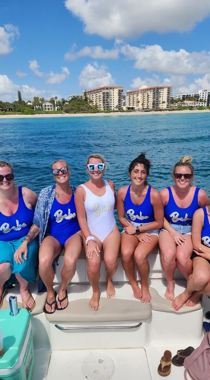 Seven women smiling on a boat in turquoise ocean during a sunny bachelorette boat party — bride in a white swimsuit and six friends in matching blue swimsuits, with sandy beach and beachfront condos on the coast behind them.