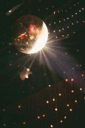 Shiny disco mirror ball hanging from a dark ceiling, brightly lit and casting star-like rays and scattered golden reflections for a retro dance-floor vibe.