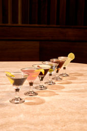 Row of six colorful martini cocktails in stemmed glasses with lemon and lime garnishes on a marble bar countertop against dark wood paneling