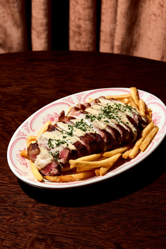 Sliced medium-rare steak drizzled with creamy sauce and chopped chives, served over crispy fries on a vintage oval plate on a dark wood table.
